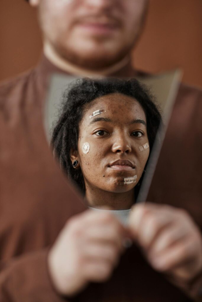 A woman with skincare patches reflecting in a broken mirror held by a bearded man.